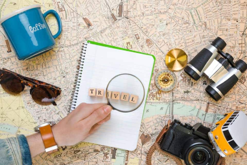 Flat lay of travel essentials: sunglasses, camera, binoculars, compass, a notepad with TRAVEL under a magnifying glass, and a coffee mug.