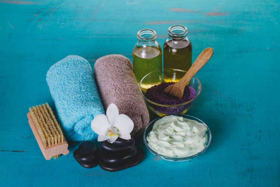 Spa essentials: rolled towels, oils, scrubs, a flower, stones, and a brush arranged on a teal wooden surface.