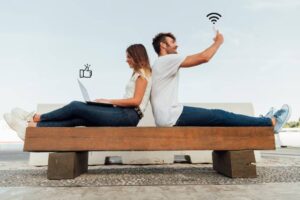 A man and woman sit back-to-back on a bench, with a laptop and phone, symbols of social media engagement and connectivity overhead.