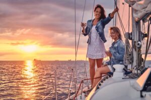 Two women on a sailboat at sunset, enjoying the ocean view with vibrant orange and purple skies reflecting on the water.