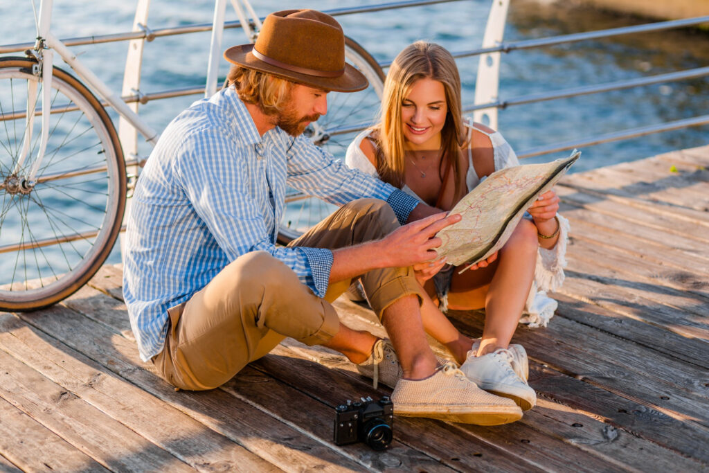 A couple sits on a wooden pier, closely examining a map with a bicycle nearby and a vintage camera at their feet.