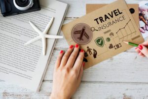 A hand with red nails holds a green pencil on a travel insurance brochure beside a camera, starfish, and a book.