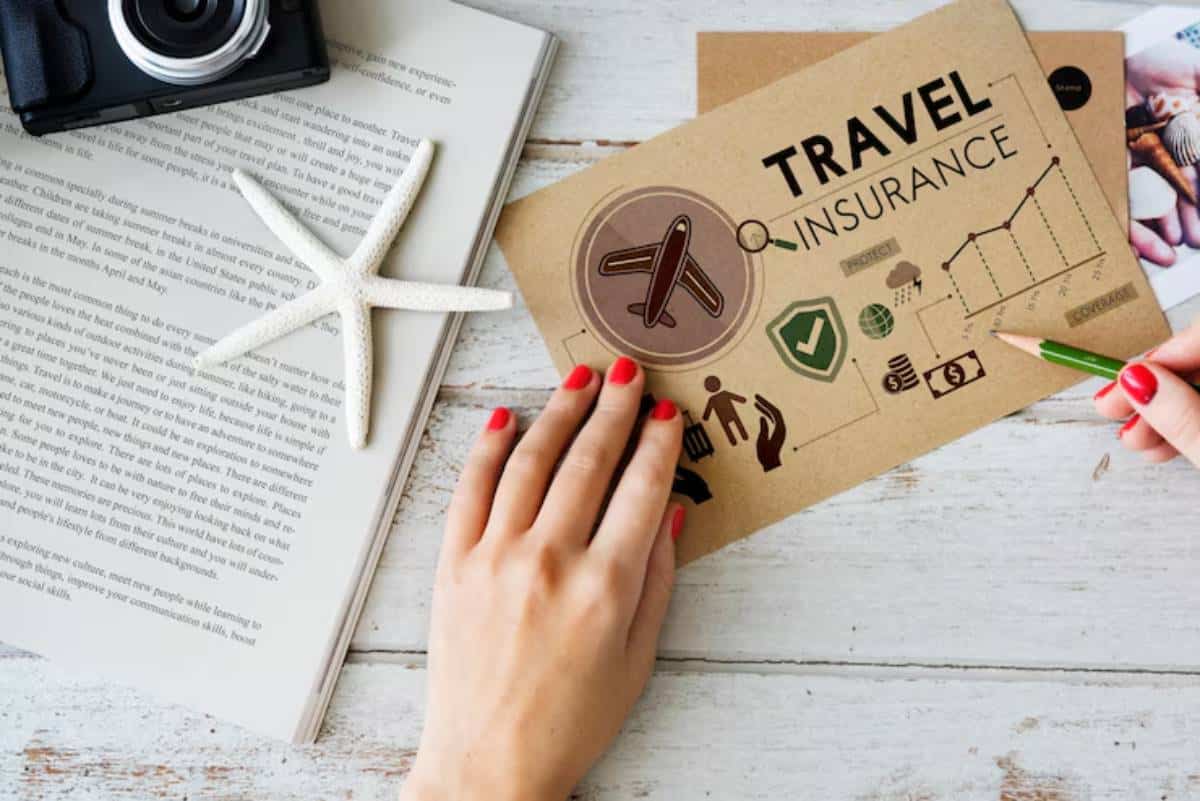 A hand with red nails holds a green pencil on a travel insurance brochure beside a camera, starfish, and a book.