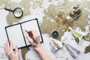 A hand writes in a notebook atop a world map surrounded by coins, a compass, paper planes, and a magnifying glass.