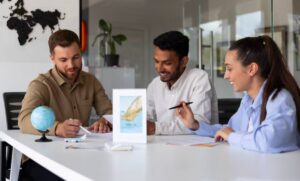 Three individuals engaged in a discussion at a modern office table, with a globe and a promotional sign visible in the background.
