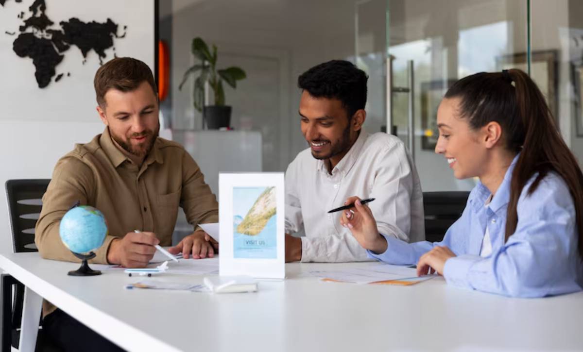 Three individuals engaged in a discussion at a modern office table, with a globe and a promotional sign visible in the background.