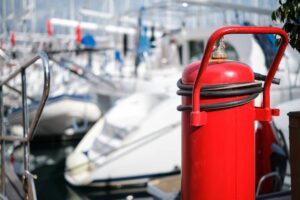 A red fire extinguisher stands near boats at a marina, emphasizing safety in a vibrant, bustling waterfront setting.
