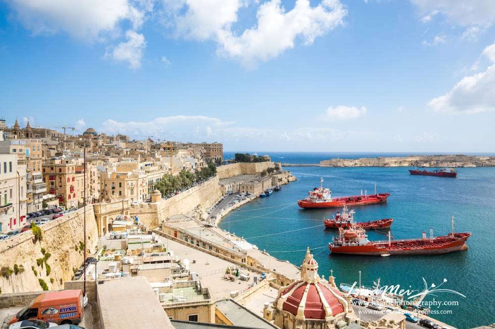 A scenic view of Valletta's harbor, featuring colorful buildings, moored red ships, and a blue sky with scattered clouds.
