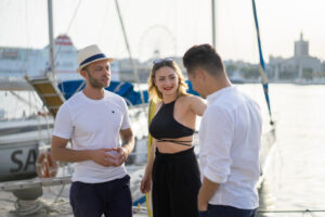 A group of three people engaged in conversation by a marina, with sailboats and a waterfront city skyline in the background.