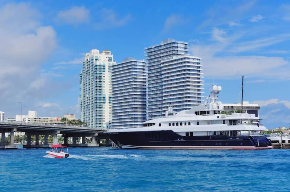 A luxurious yacht navigates the turquoise waters near modern high-rise buildings under a bright blue sky in a coastal city.