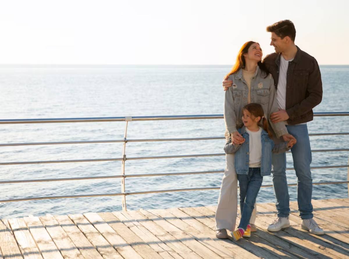 A family stands together on a wooden deck by the sea, enjoying a sunny day with the ocean in the background.