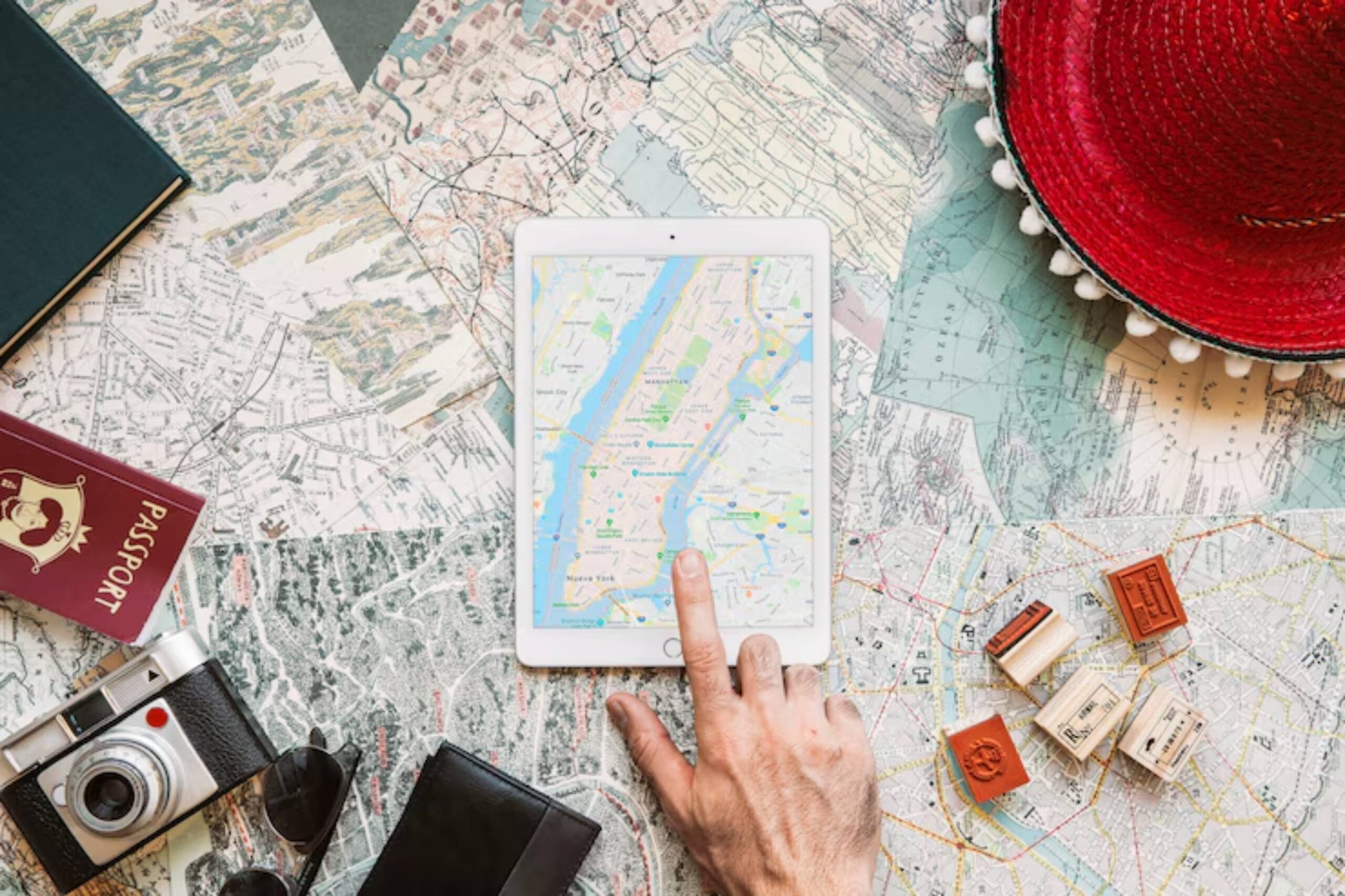 A hand points at a digital map on a tablet surrounded by travel-related items like maps, a camera, a passport, and a red hat.