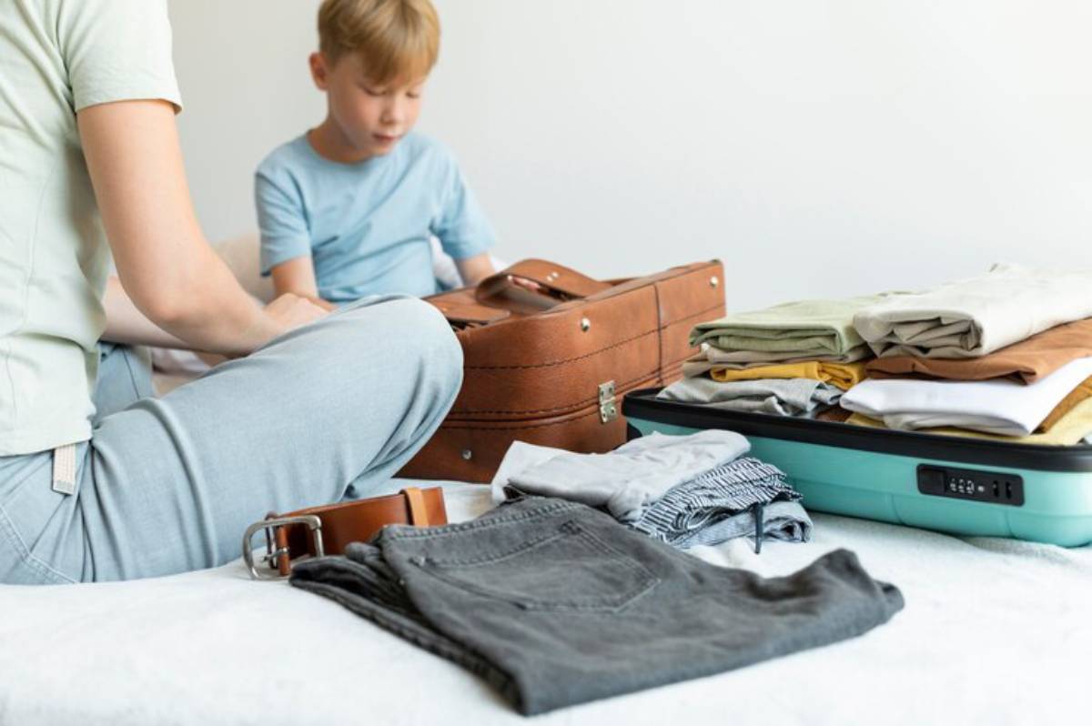 A person sits on a bed surrounded by neatly folded clothes and a brown suitcase, preparing for travel.
