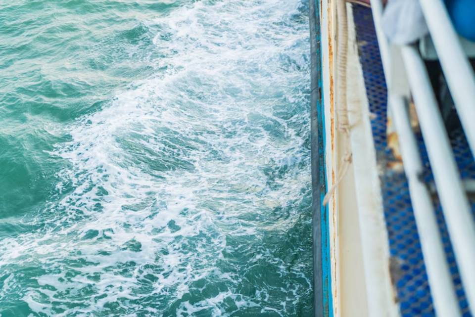 Waves ripple against the side of a ferry, with the turquoise water contrasting against the white foam and ferry's rusted edges.