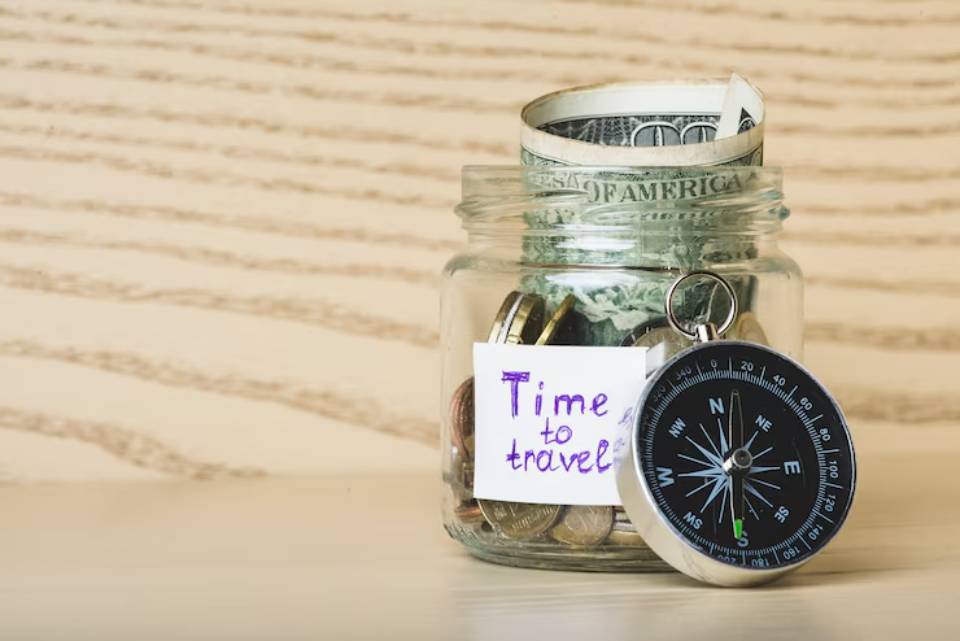 A jar filled with coins and a rolled dollar bill, labeled Time to travel,next to a compass on a wooden background.