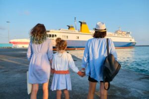 Two women and a girl hold hands, facing a large ferry at a sunny port, ready for an adventure. Their outfits are casual and beachy.