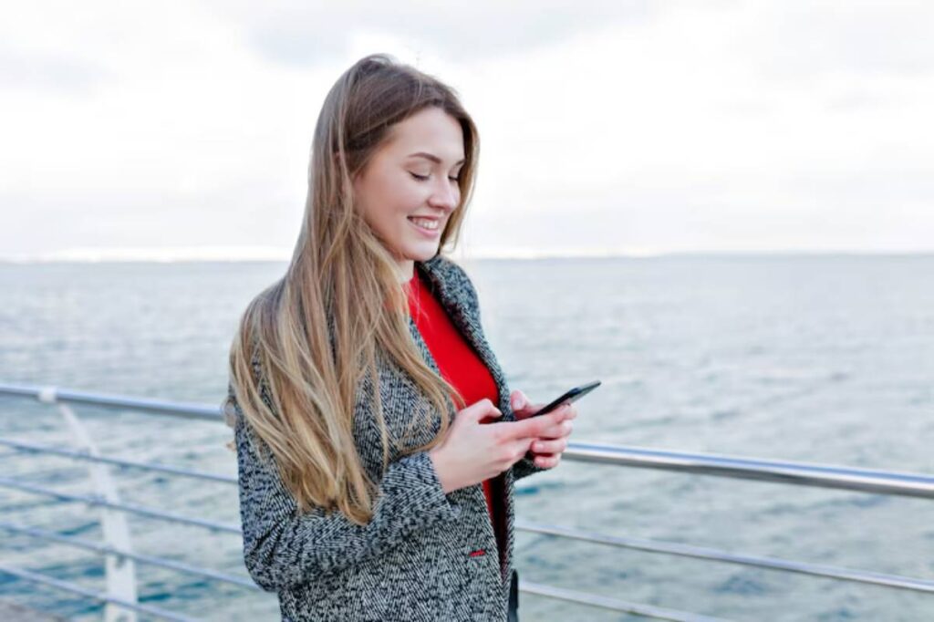 A woman in a grey coat and red top, standing by a railing overlooking the water, is focused on her smartphone.