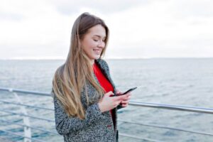A woman in a grey coat and red top, standing by a railing overlooking the water, is focused on her smartphone.