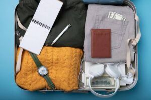A neatly packed suitcase containing clothes, a notebook, a pen, a passport, a watch, headphones, and cash, against a blue background.